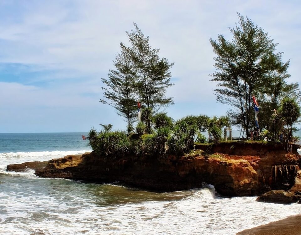 Rocky island covered with palm trees extends into calm blue ocean under a clear sky with gentle waves.