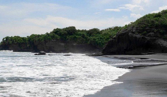 Black-sand beach with foamy waves and tree-covered cliffs in the distance.