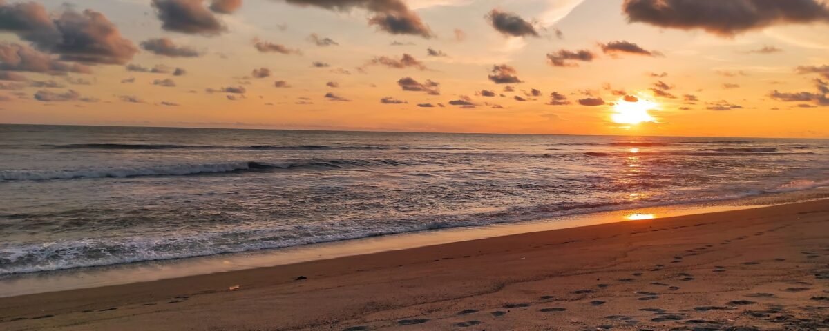 Sunset over the ocean with an orange sky and gentle waves; footprints in the wet sand in the foreground.