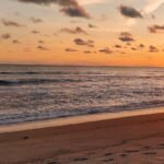 Sunset over the ocean with an orange sky and gentle waves; footprints in the wet sand in the foreground.