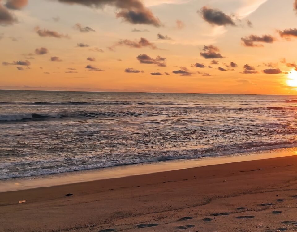 Sunset over the ocean with an orange sky and gentle waves; footprints in the wet sand in the foreground.