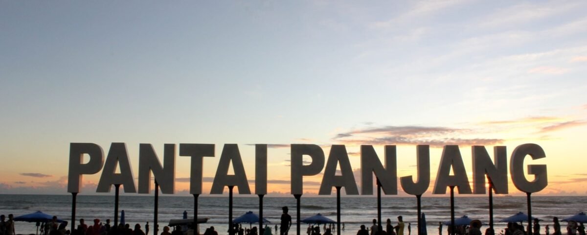 Large sign reading 'PANTAI PANGJANG' on a beach at sunset with people and umbrellas remaining in the foreground.
