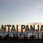 Large sign reading 'PANTAI PANGJANG' on a beach at sunset with people and umbrellas remaining in the foreground.