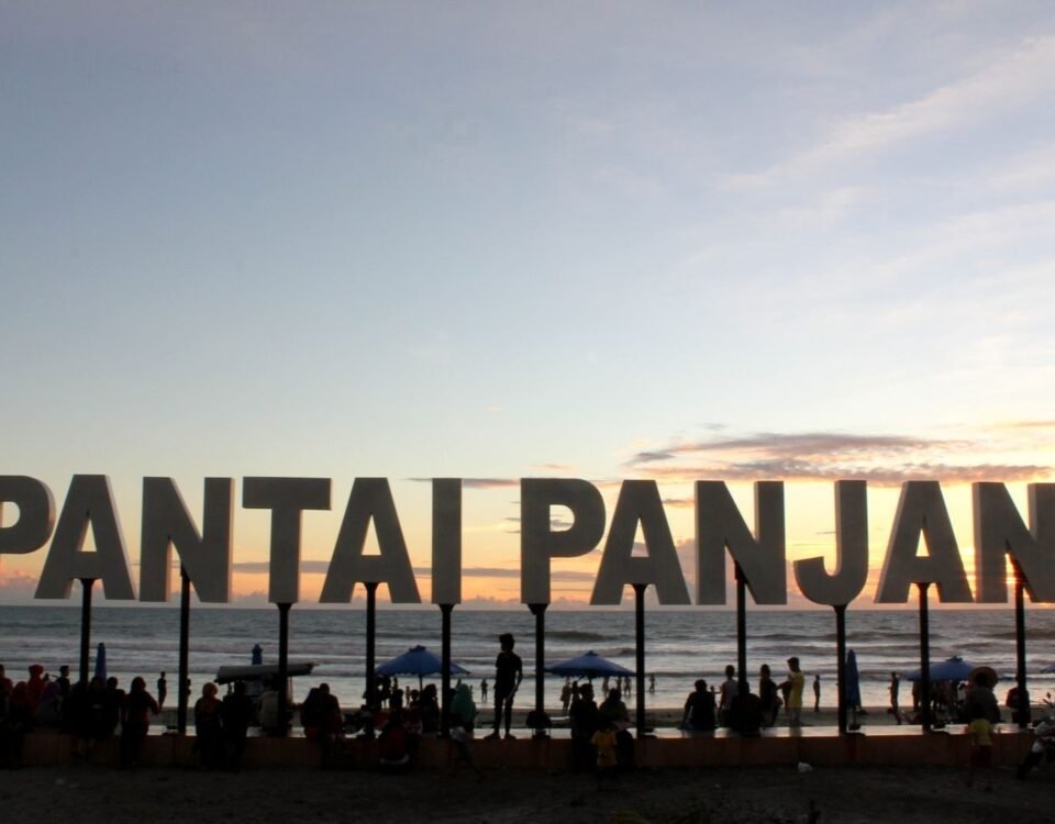 Large sign reading 'PANTAI PANGJANG' on a beach at sunset with people and umbrellas remaining in the foreground.