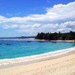 Sunny tropical beach with clear blue water, gentle waves, and anchored boats near the shore; tree-lined coastline in the distance.
