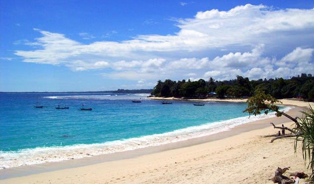 Sunny tropical beach with clear blue water, gentle waves, and anchored boats near the shore; tree-lined coastline in the distance.