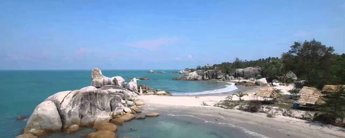 Tropical beach scene with large granite boulders along a turquoise sea and a sandy shore under a clear blue sky, with trees on the right.