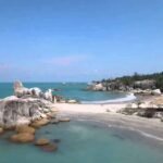 Tropical beach scene with large granite boulders along a turquoise sea and a sandy shore under a clear blue sky, with trees on the right.