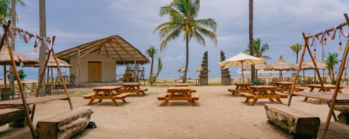 Beachside cafe area with wooden picnic tables, swings, palm trees, and a thatched hut overlooking the ocean.