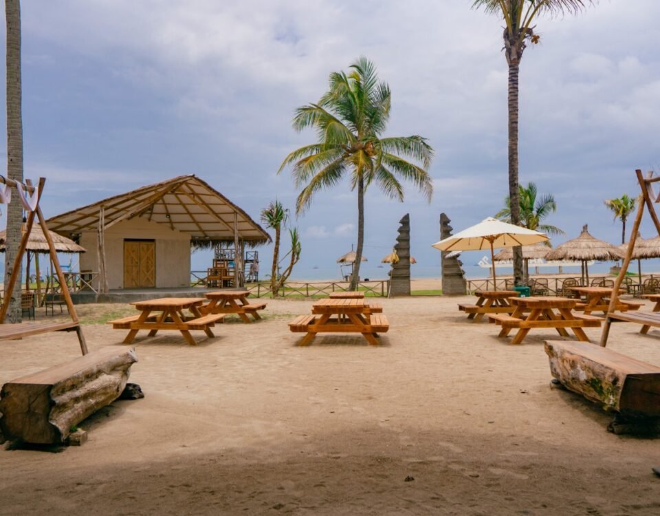 Beachside cafe area with wooden picnic tables, swings, palm trees, and a thatched hut overlooking the ocean.