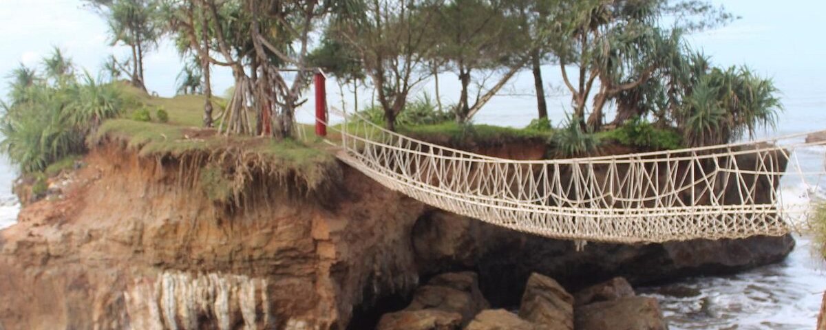 Rope suspension bridge spanning from rocky cliff to a small tree-covered island by the sea.