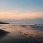 Sunset over a calm beach with palm trees on the left and wet sand reflecting pink sky.
