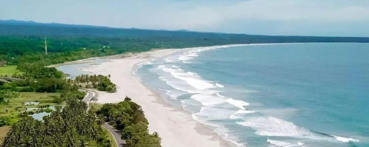 Aerial view of a tropical coastline with a long white-sand beach, turquoise water, and a tree-lined road along the shore.