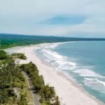 Aerial view of a tropical coastline with a long white-sand beach, turquoise water, and a tree-lined road along the shore.