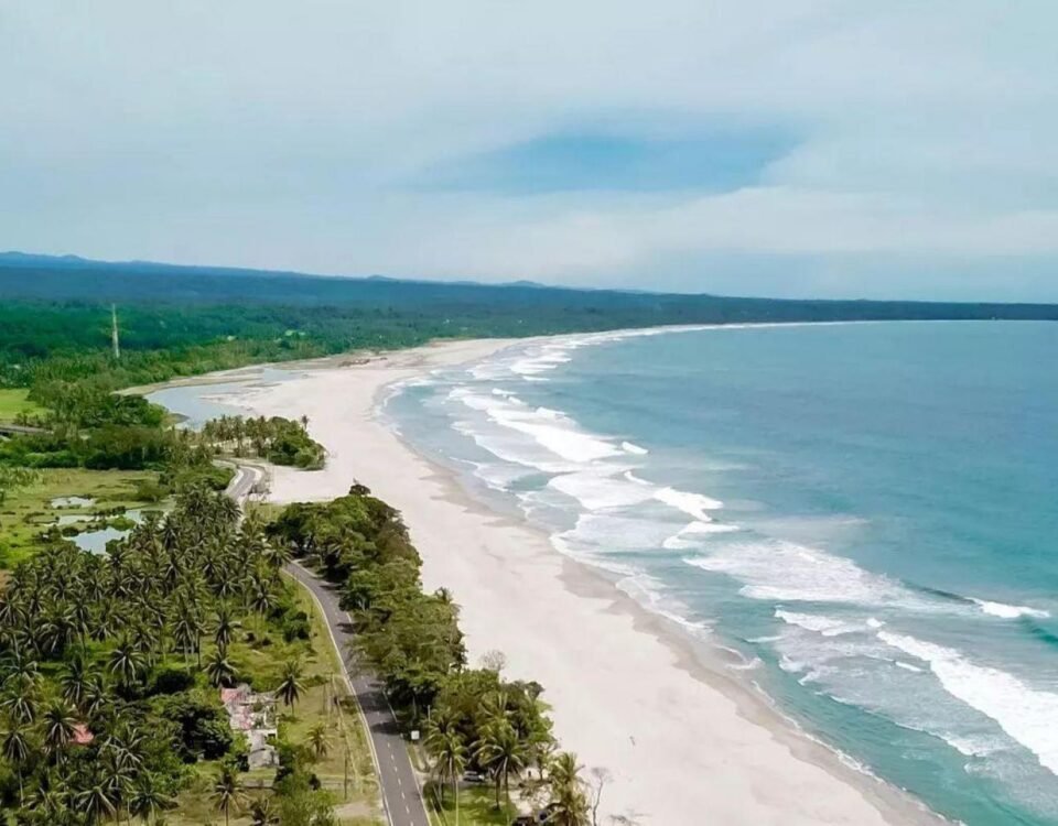 Aerial view of a tropical coastline with a long white-sand beach, turquoise water, and a tree-lined road along the shore.