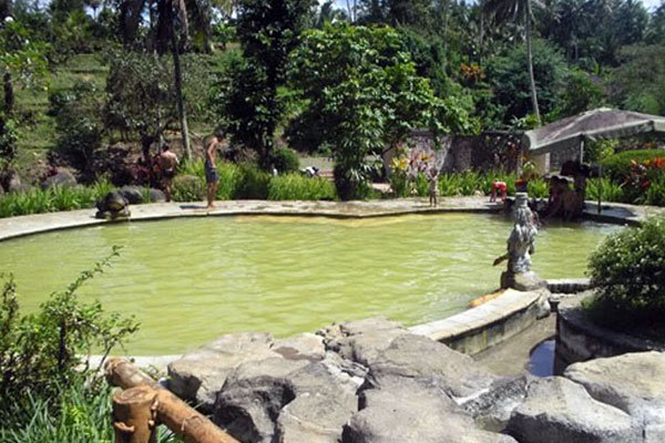 Outdoor garden pool with stone edge and greenish water, people relaxing by the rim amidst tropical trees and rocks