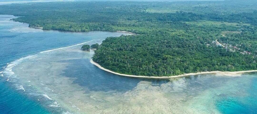 Aerial view of a tropical island with dense green forest, white-sand beaches, and a turquoise lagoon encircled by a coral reef.