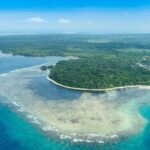 Aerial view of a tropical island with dense green forest, white-sand beaches, and a turquoise lagoon encircled by a coral reef.