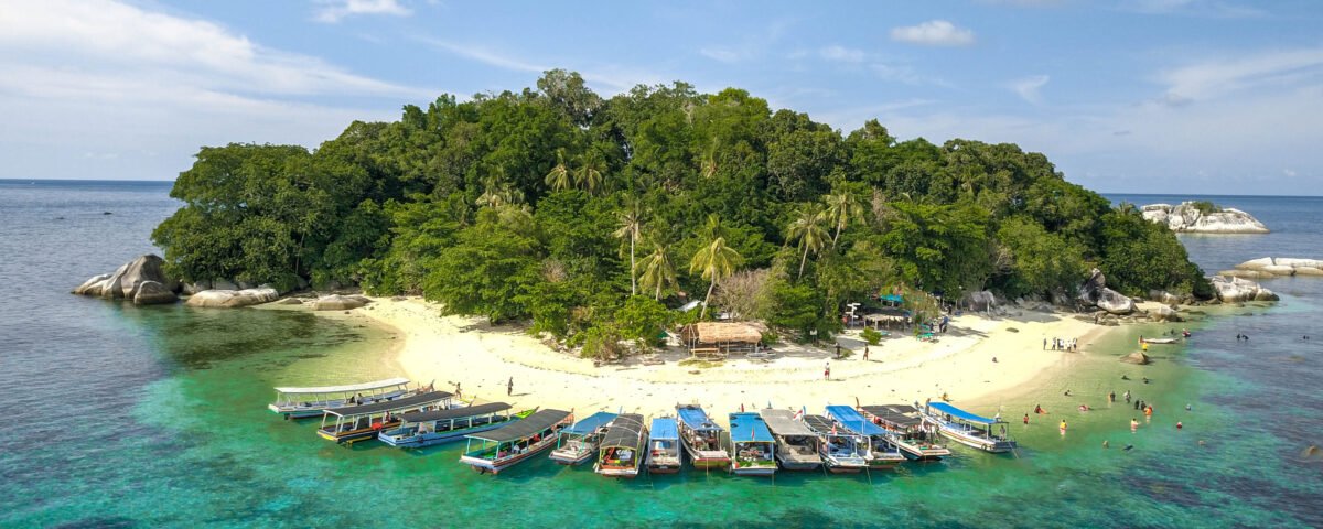 A small tropical island with a sandy beach, dense green trees, and boats docked along the shore.
