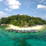 A small tropical island with a sandy beach, dense green trees, and boats docked along the shore.