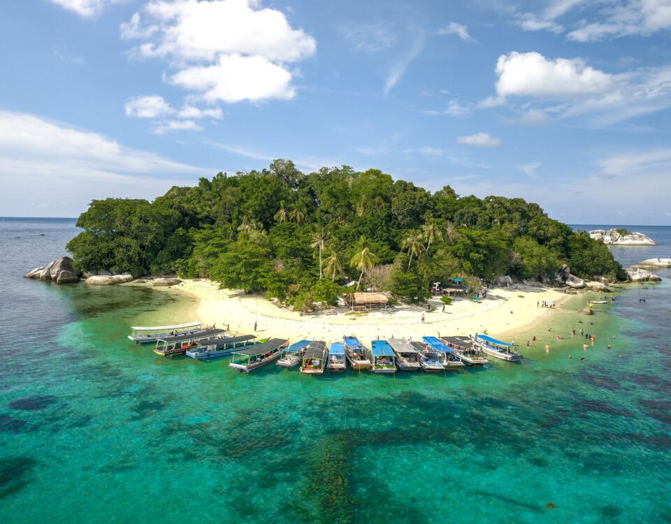 A small tropical island with a sandy beach, dense green trees, and boats docked along the shore.