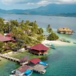 Tropical shoreline with palm trees, thatched-roof huts on stilts, and turquoise water; boats docked along a wooden pier.