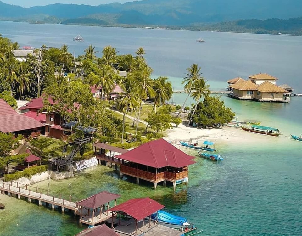 Tropical shoreline with palm trees, thatched-roof huts on stilts, and turquoise water; boats docked along a wooden pier.