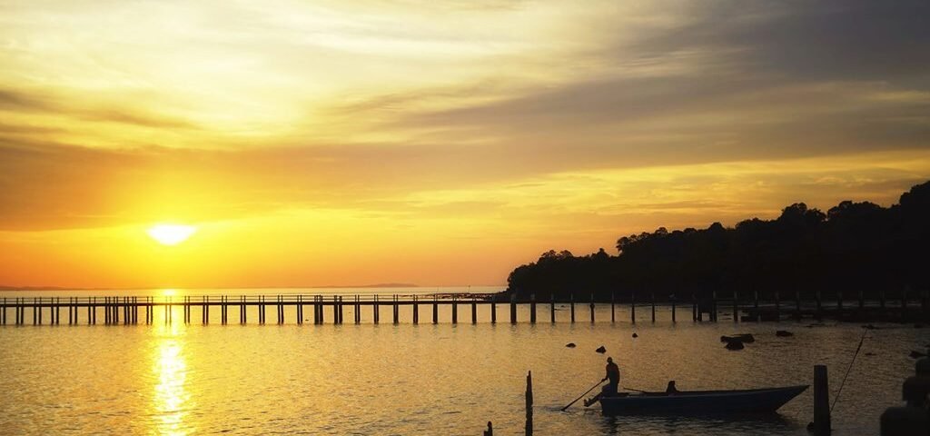 Golden sunset over a calm bay with a long wooden pier reaching toward the horizon and a small boat gliding nearby. The scene is reflected in the water.