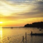 Golden sunset over a calm bay with a long wooden pier reaching toward the horizon and a small boat gliding nearby. The scene is reflected in the water.