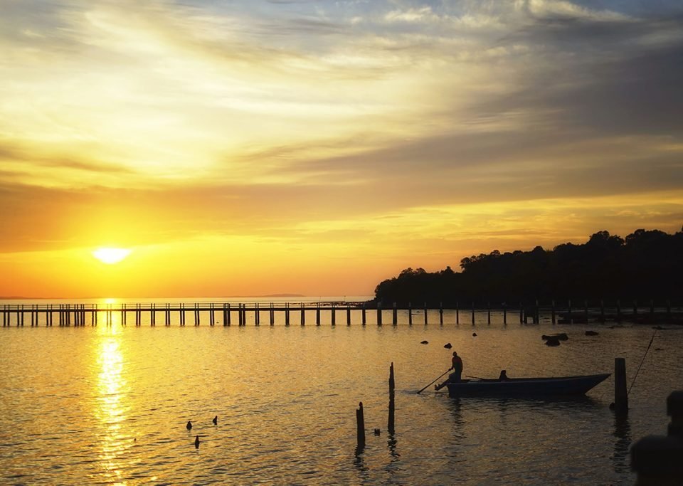 Golden sunset over a calm bay with a long wooden pier reaching toward the horizon and a small boat gliding nearby. The scene is reflected in the water.