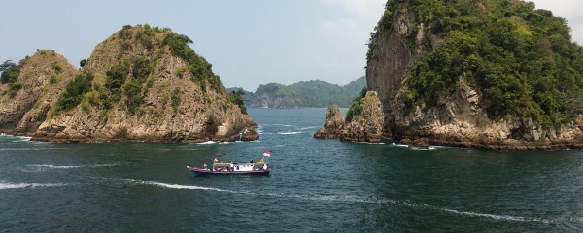 Boat with Indonesian flag sails through a narrow channel between two rocky, green-covered islets in calm blue water under a clear sky.