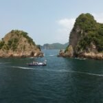 Boat with Indonesian flag sails through a narrow channel between two rocky, green-covered islets in calm blue water under a clear sky.