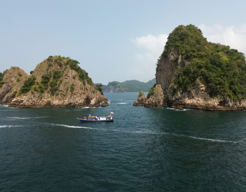 Boat with Indonesian flag sails through a narrow channel between two rocky, green-covered islets in calm blue water under a clear sky.