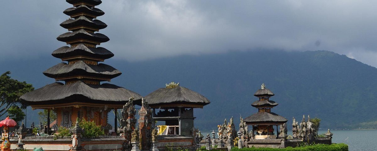 Stone and thatch-roofed temple complex on a lake shore with a tall multi-tiered pagoda and smaller shrines, mountains in the background