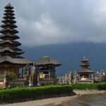 Stone and thatch-roofed temple complex on a lake shore with a tall multi-tiered pagoda and smaller shrines, mountains in the background