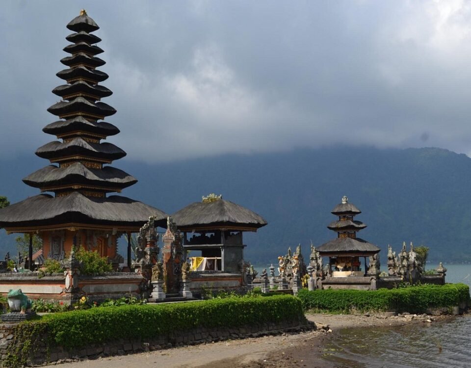 Stone and thatch-roofed temple complex on a lake shore with a tall multi-tiered pagoda and smaller shrines, mountains in the background