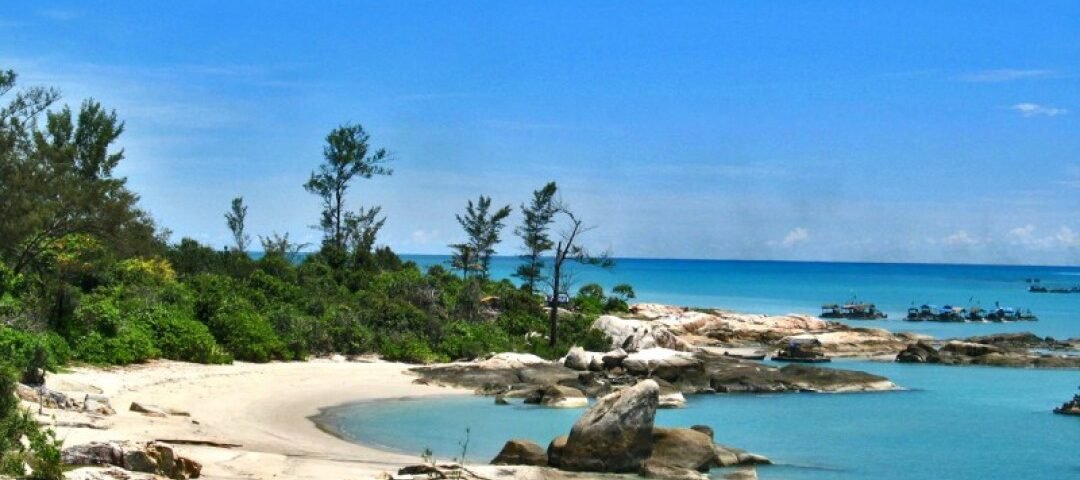 Tropical beach with pale sand, turquoise water, and large rocks along a rocky shoreline under a bright blue sky.