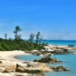 Tropical beach with pale sand, turquoise water, and large rocks along a rocky shoreline under a bright blue sky.