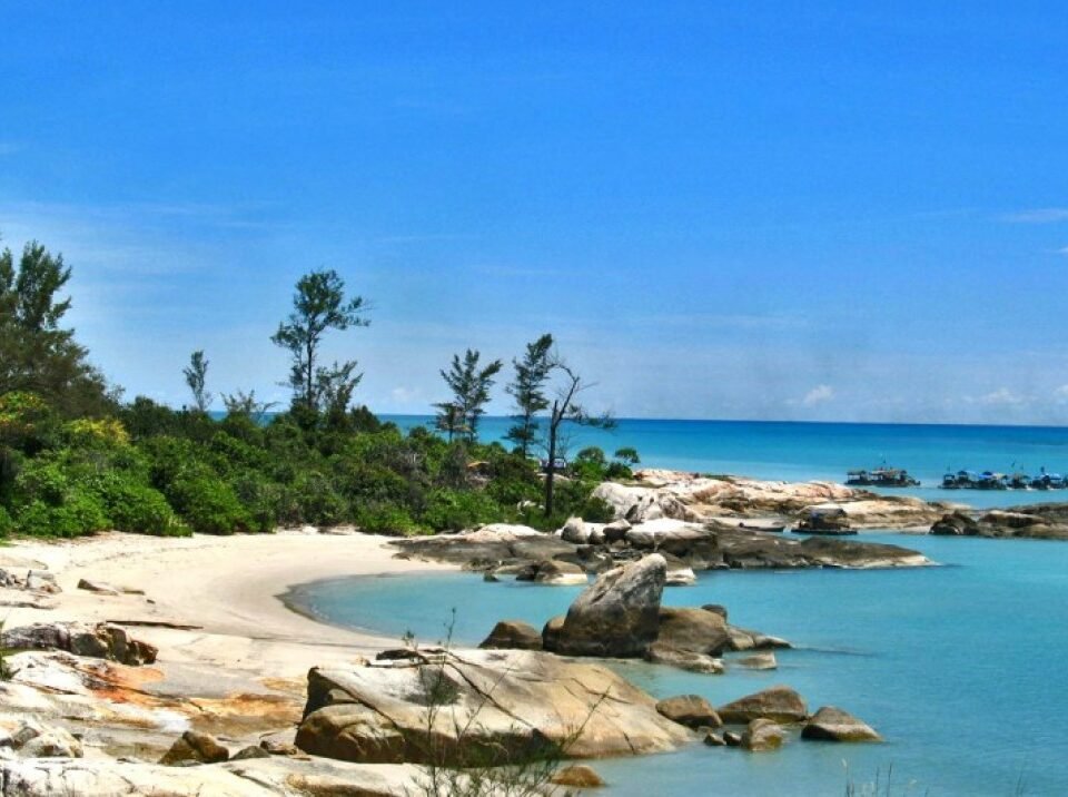 Tropical beach with pale sand, turquoise water, and large rocks along a rocky shoreline under a bright blue sky.
