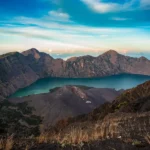 Turquoise crater lake surrounded by rugged volcanic peaks under a blue sky with wispy clouds.