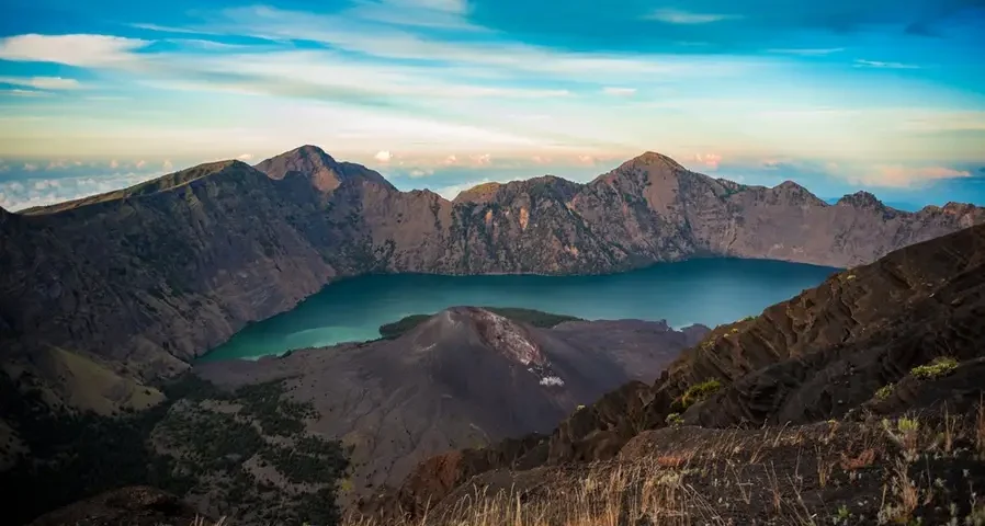 Turquoise crater lake surrounded by rugged volcanic peaks under a blue sky with wispy clouds.