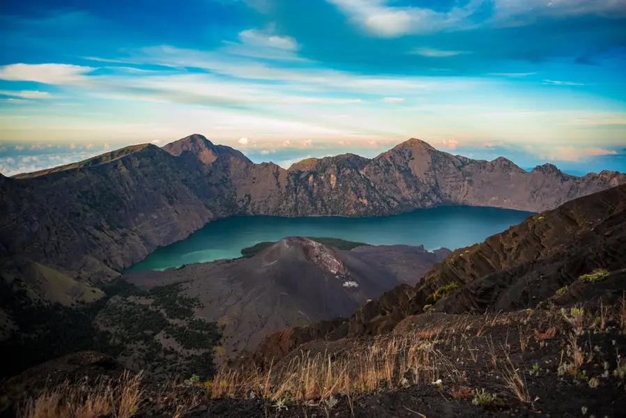 Turquoise crater lake surrounded by rugged volcanic peaks under a blue sky with wispy clouds.