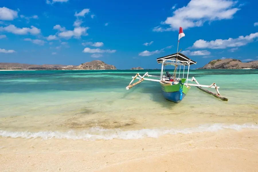Colorful wooden outrigger boat anchored on a sandy tropical beach with turquoise water and a bright blue sky.