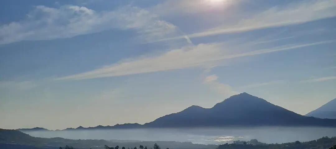 Sun over a blue sky above a misty valley with distant mountains and a green foreground hillside.