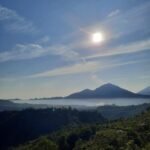 Sun over a blue sky above a misty valley with distant mountains and a green foreground hillside.