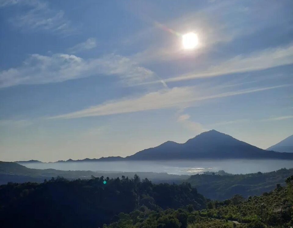 Sun over a blue sky above a misty valley with distant mountains and a green foreground hillside.