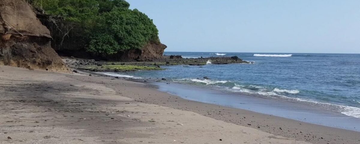 Sandy beach with a rocky, tree-covered headland on the left and calm blue ocean to the right under a clear sky.