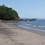 Sandy beach with a rocky, tree-covered headland on the left and calm blue ocean to the right under a clear sky.