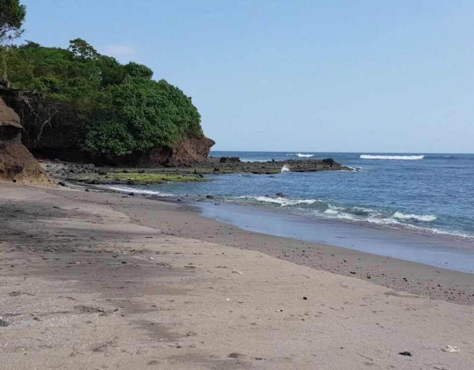 Sandy beach with a rocky, tree-covered headland on the left and calm blue ocean to the right under a clear sky.