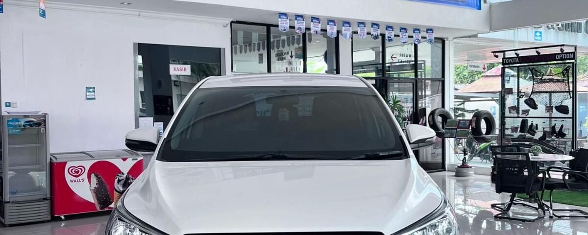 Front view of a white Toyota SUV on a showroom floor, bold black grille and emblem visible, with banners and glass doors in the background.